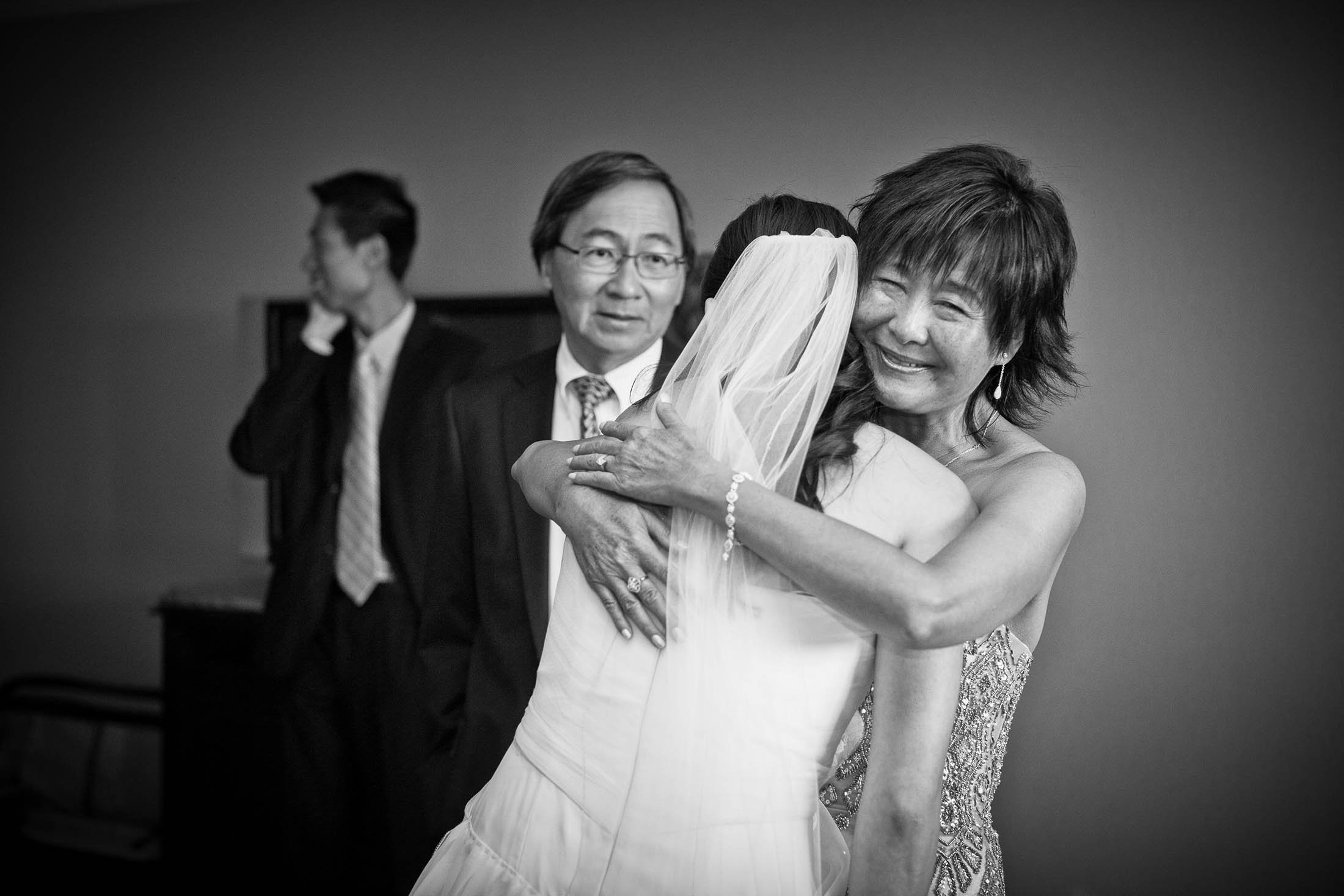 A smiling woman hugs a bride in a wedding dress and veil. Two men in suits stand in the background; one looks away while the other faces forward. The scene is in black and white.