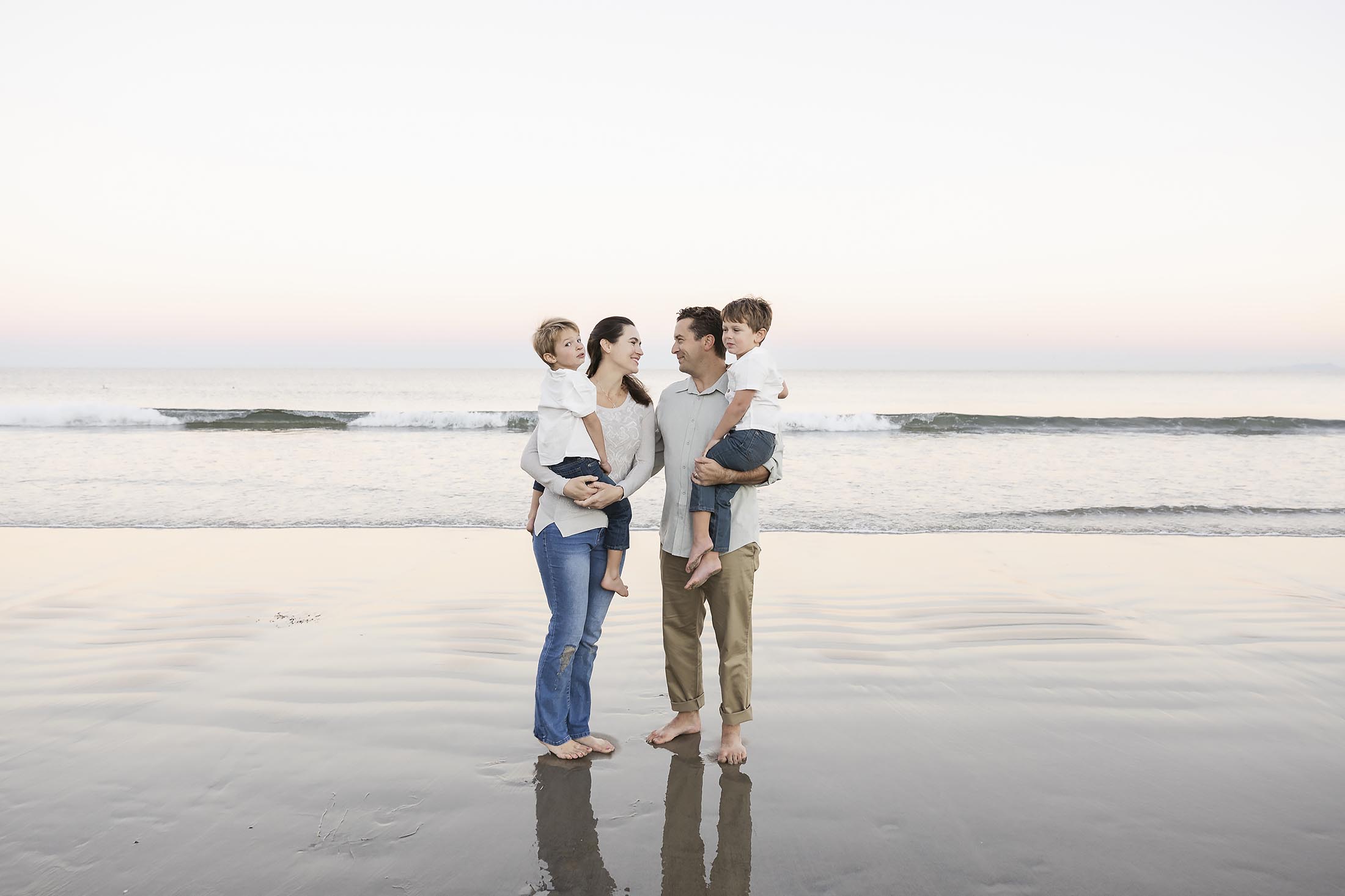 A family of four stands barefoot on a beach, with gentle waves in the background. Captured by a talented family photographer, the parents hold their two young children—all smiling and savoring a peaceful moment together by the sea.