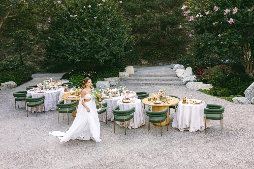 A woman in a white dress walks past elegantly set tables with green chairs and floral centerpieces on a stone patio at the Japanese Friendship Garden.