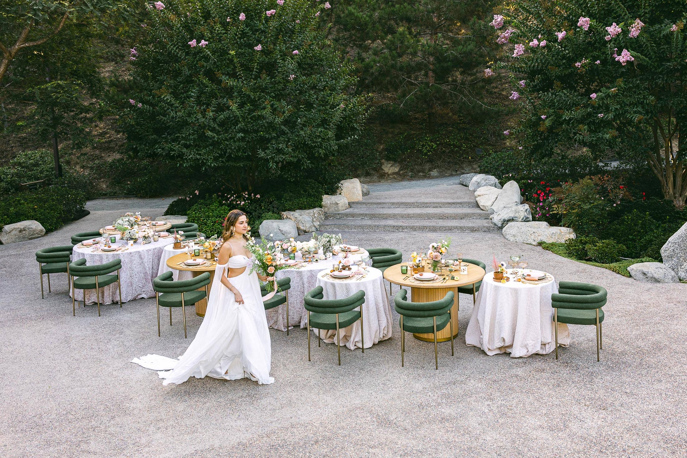 A woman in a white dress walks past elegantly set tables with green chairs and floral centerpieces on a stone patio surrounded by greenery.