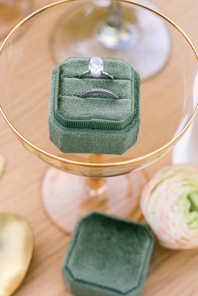 A diamond engagement ring and a matching wedding band displayed in an open, green velvet ring box atop a gold-rimmed glass. A closed box and a decorative flower are nearby, placed on a wooden table.