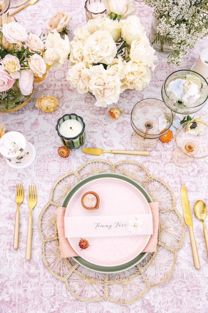 Elegant table setting with a pink and green plate, gold cutlery, and a woven placemat. Candles and floral arrangements with white and pink roses are placed on a patterned tablecloth. A name card reads "Tommy Fair.