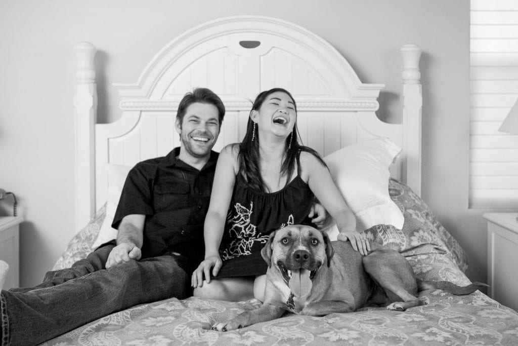 A black and white image captures the essence of San Diego engagement photosโ a couple sitting on a bed with their large dog, laughing and relaxed. The dog sits contently between them in the airy room, complete with a white headboard, embodying joy and tranquility.