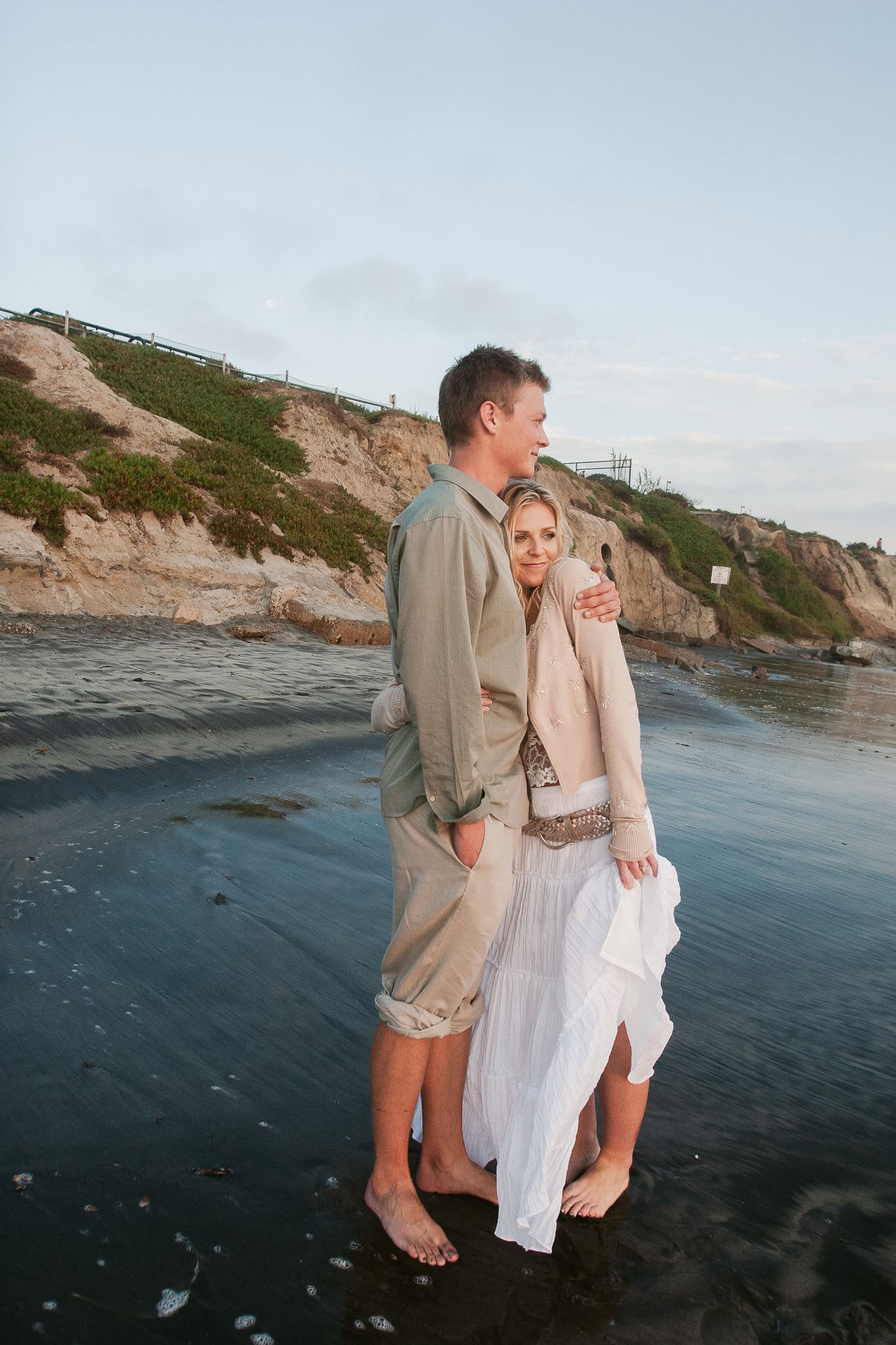 A couple stands barefoot on a San Diego beach, with the man hugging the woman. They are dressed in casual, light-colored clothes, perfect for engagement photos. The beach has wet sand and grassy cliffs in the background, while the sky is cloudy with soft lighting.