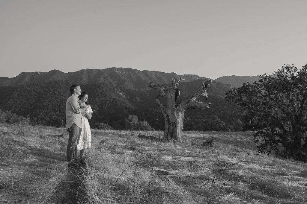 A couple stands in a grassy, open field, gazing thoughtfully at the scenic mountains near San Diego. A large, bare tree is nearby, adding contrast to the serene landscape under a clear sky. The black and white image captures an enchanting moment akin to engagement photos.
