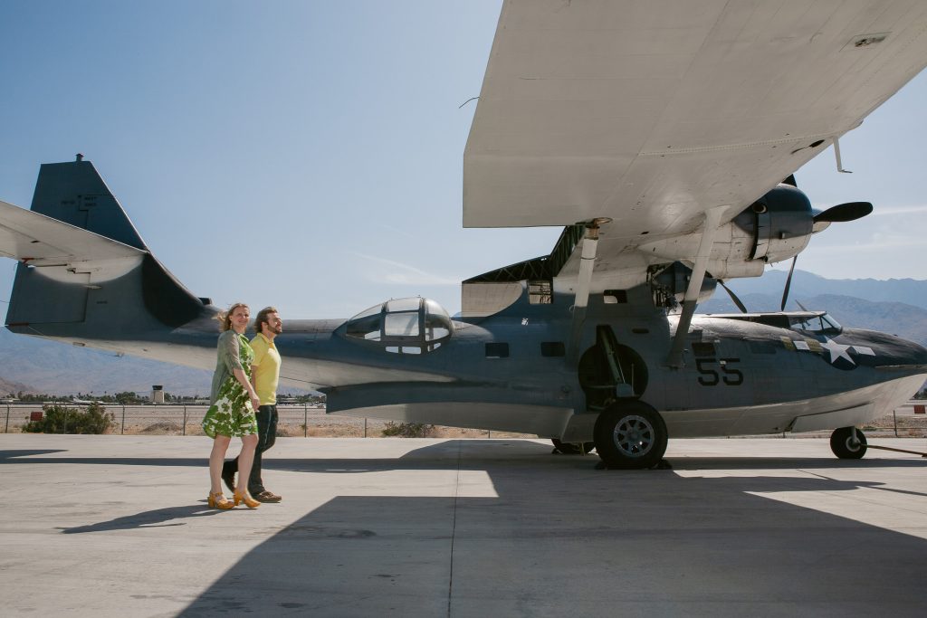 Two people stand beside a vintage seaplane with large wings and propellers, perfect for their San Diego engagement photos. The plane, painted in grey with the number "55," is parked on a sunny tarmac, with majestic mountains visible in the background.