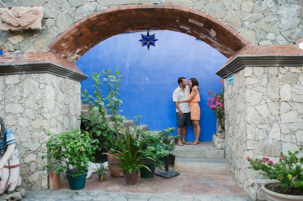 A couple stands under a stone archway in front of a vibrant blue wall in their San Diego engagement photos. The woman wears an orange dress, and the man is in a light shirt and shorts. Potted plants and stone decorations surround them, creating a charming and cozy atmosphere.