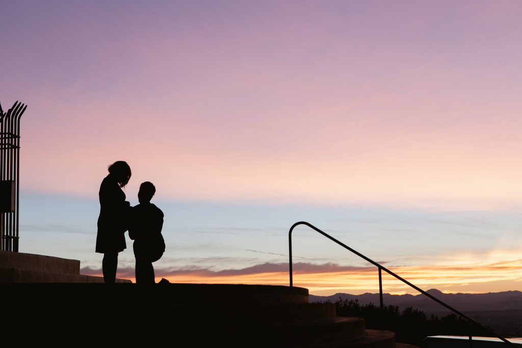 Silhouette of two people on steps at sunset in a San Diego engagement photo. One is kneeling, and the other stands, crafting a romantic atmosphere against a colorful sky with soft orange, pink, and purple hues. A railing and part of a structure are visible.