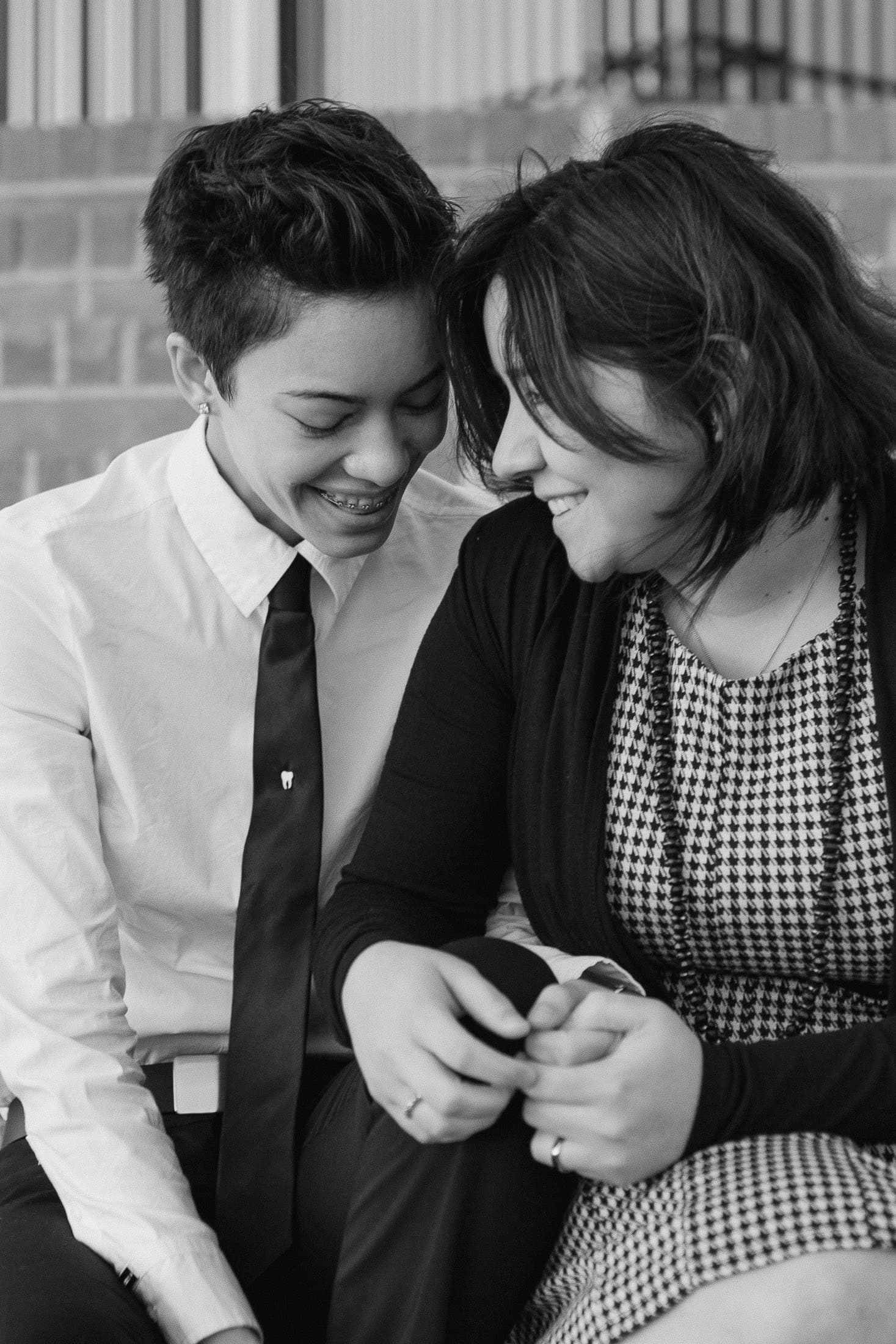 A black and white photo captures a joyful, intimate moment as two people sit closely together on steps. Dressed in a white shirt and tie, and a patterned dress with a cardigan, they smile warmly. This tender San Diego engagement photo embodies love's timeless essence.