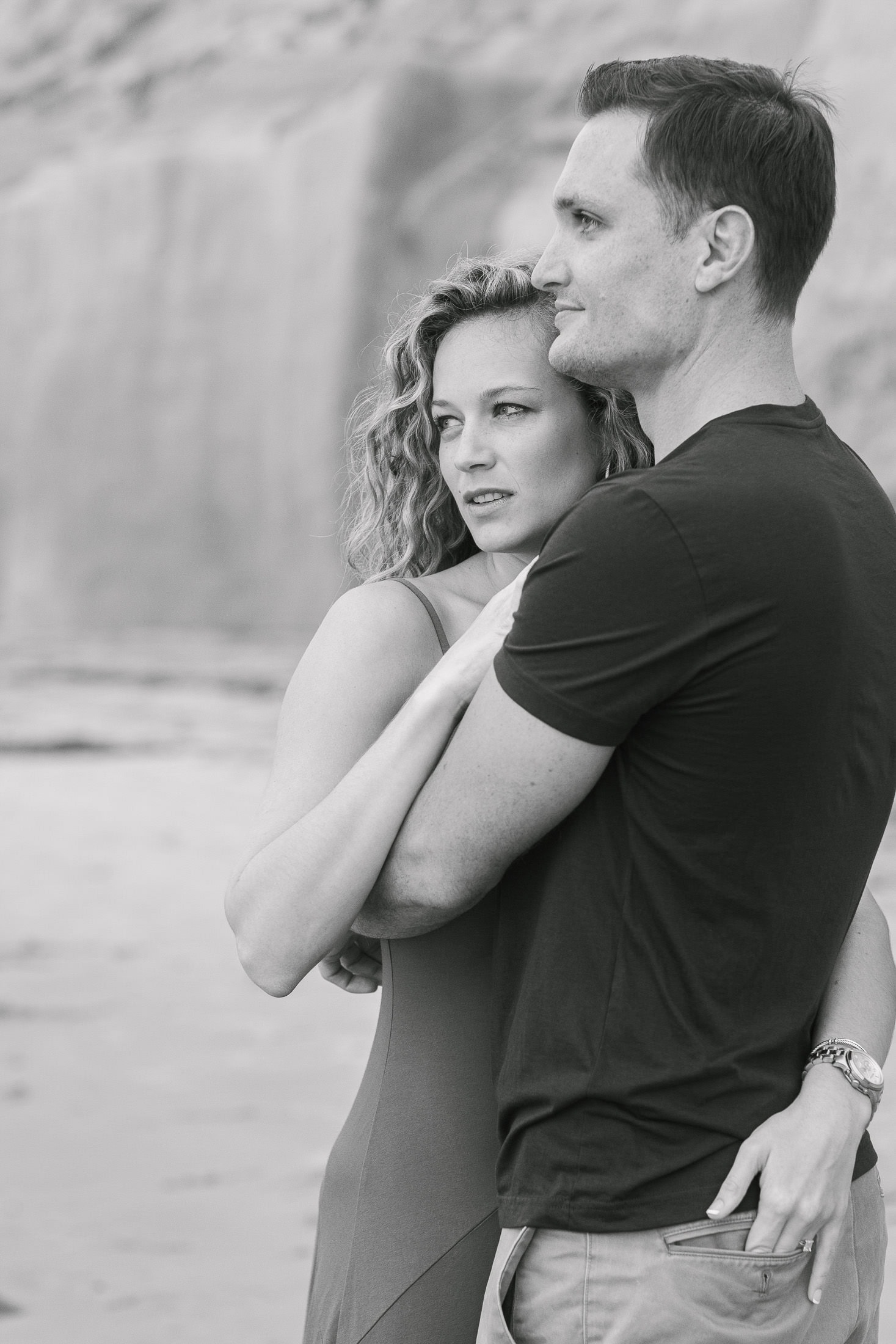 A black and white image captures a couple standing on a San Diego beach. The woman, with curly hair, leans against the man's chest as he embraces her while gazing into the distance. A sandy cliff forms the backdrop in this tender engagement photo.