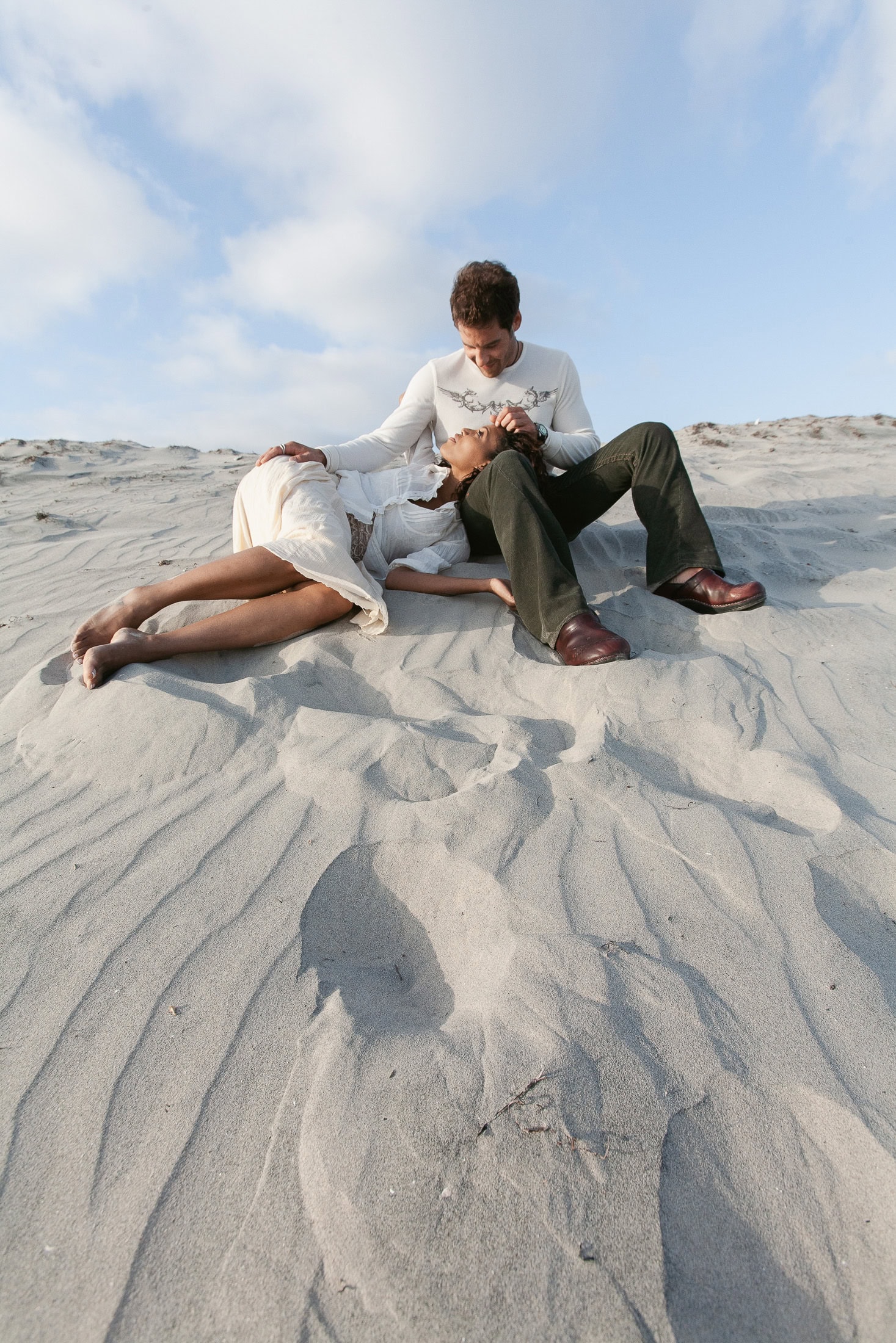A couple sits and lies on the sandy dunes under the blue San Diego sky, capturing intimate engagement photos. The woman rests her head in the man's lap while he looks down at her with a relaxed and content expression. They are dressed in light, casual clothing, perfectly fitting the serene setting.