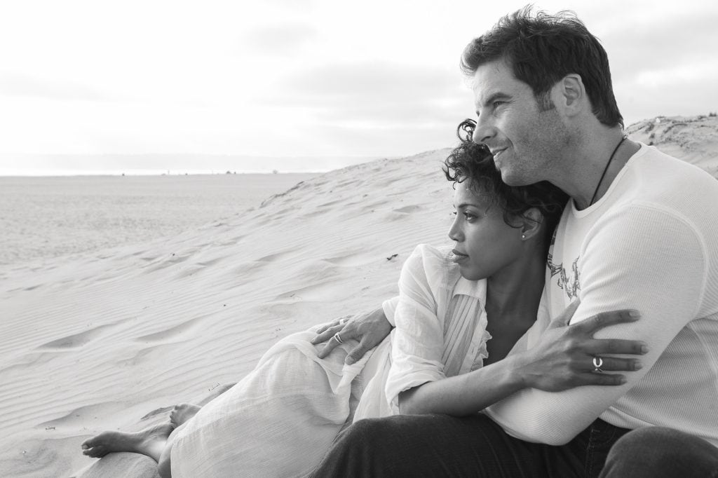 A couple sits together on a sandy beach, embracing while gazing at the horizon. The woman leans on the man's chest, their San Diego engagement photos capturing this timeless, serene scene in black and white.