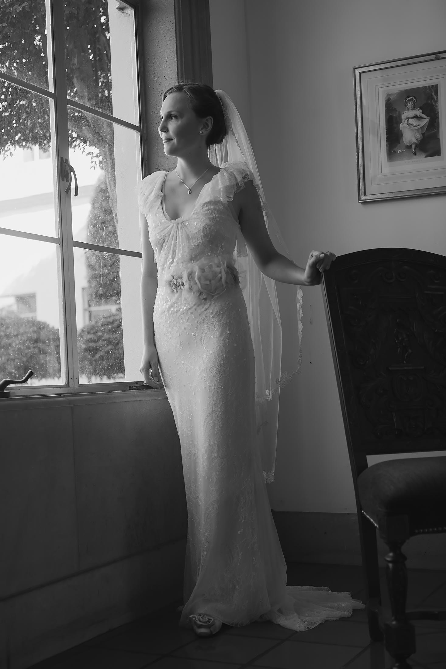 A bride in a lace wedding dress and veil stands by a large window, looking outside. She has her hand on a wooden chair. The room is decorated with a framed picture on the wall. The scene is in black and white.