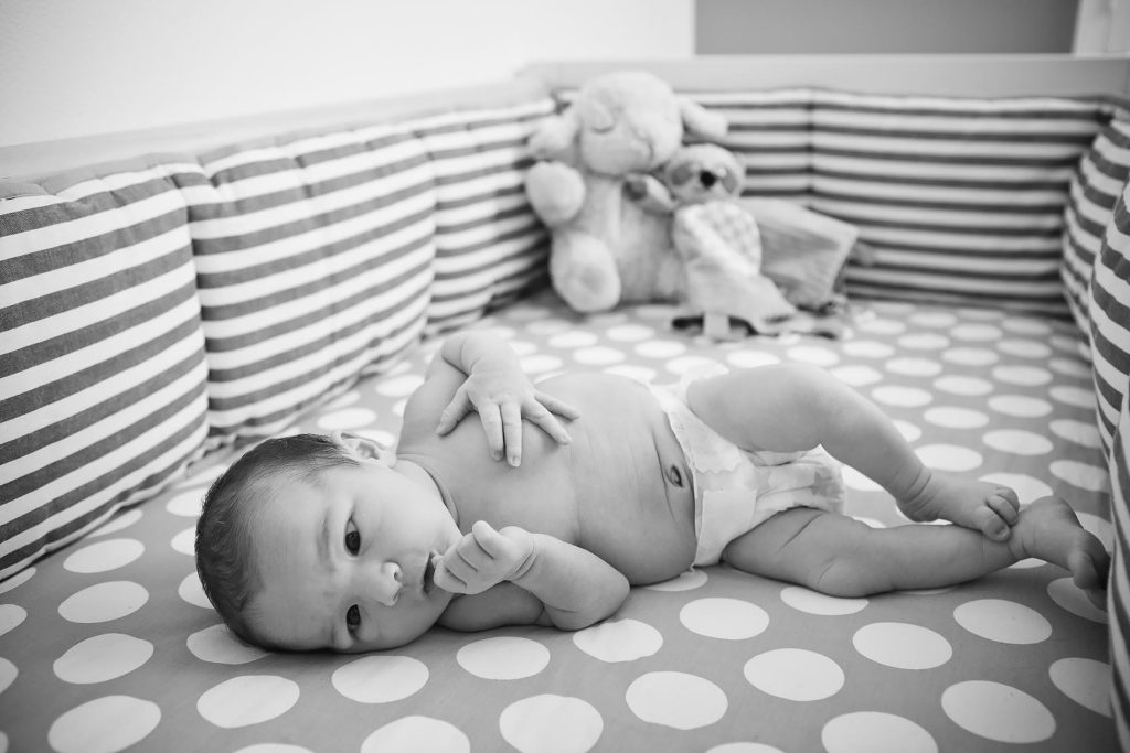 A black and white image of a baby lying on its side in a crib with a polka dot mattress. The crib has striped bumpers. In the background, there's a stuffed animal. The baby is wearing a diaper and resting one hand near its mouth.