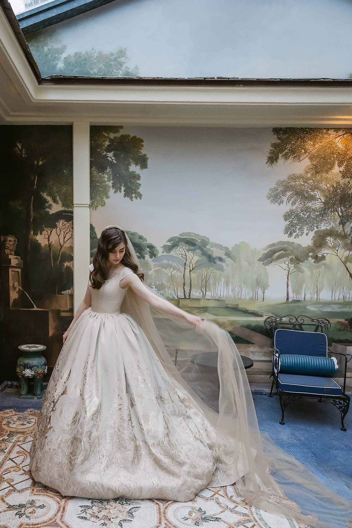 About: A woman in an elegant, gold-embroidered ball gown with a long tulle veil stands in a sunlit room with a mural of trees and a blue bench, gracefully holding her veil out to the side.