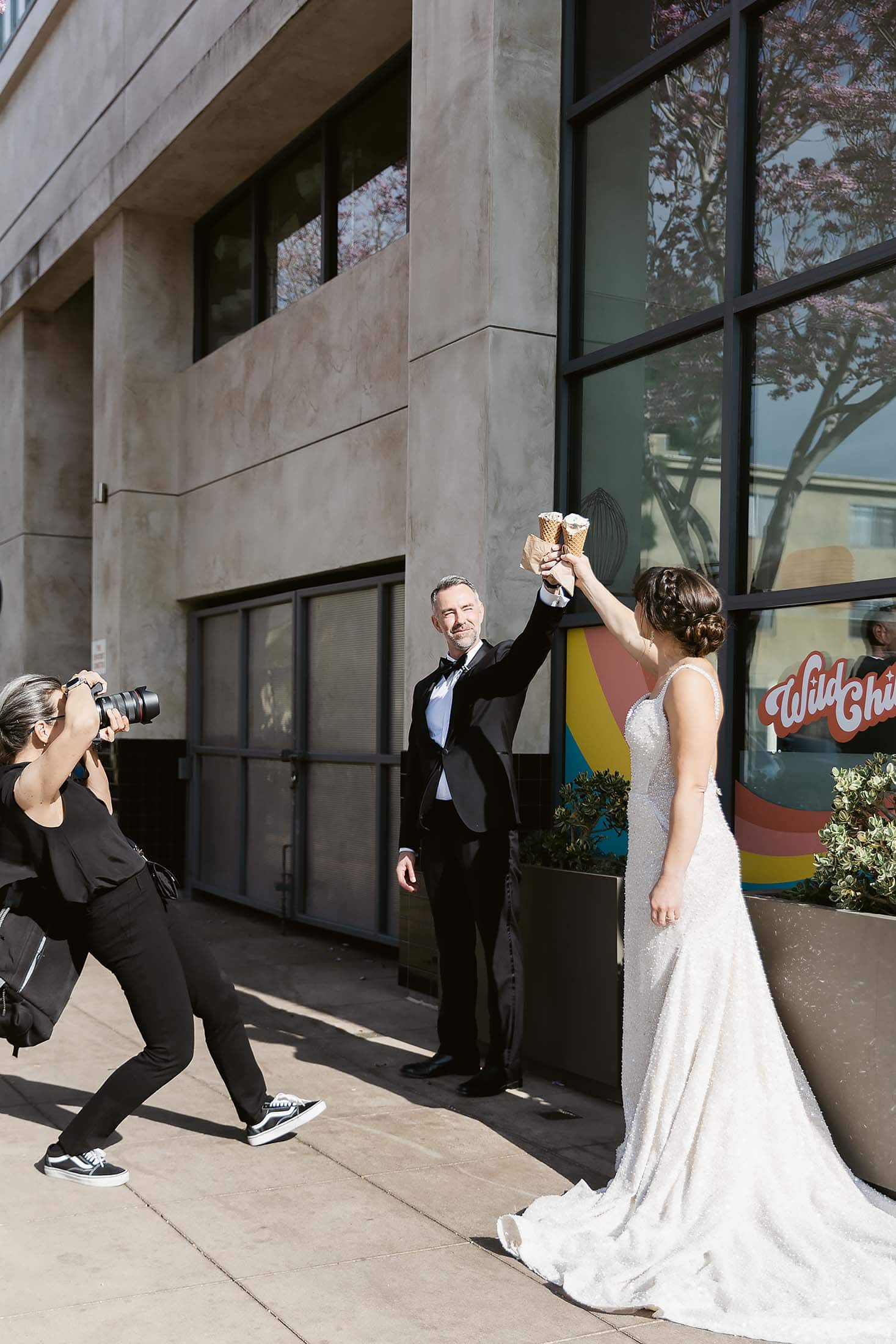 A bride and groom in formal attire toast with ice cream cones outside a modern building, while Jennifer Dery captures the moment.