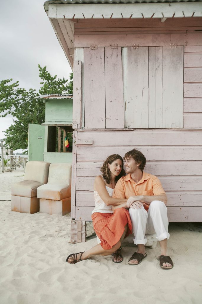 Kuyaba Hotel, Negril, Jamaica: A couple sits together on the sand, smiling and leaning close in front of a pink wooden beach hut, with mismatched chairs and green building in the background. Both are dressed in casual, summery clothes.