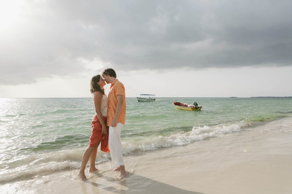 Negril, Jamaica: A couple stands barefoot on a sandy beach at the water’s edge, embracing and sharing a kiss. The sky is overcast, and two small boats float on the turquoise sea in the background.