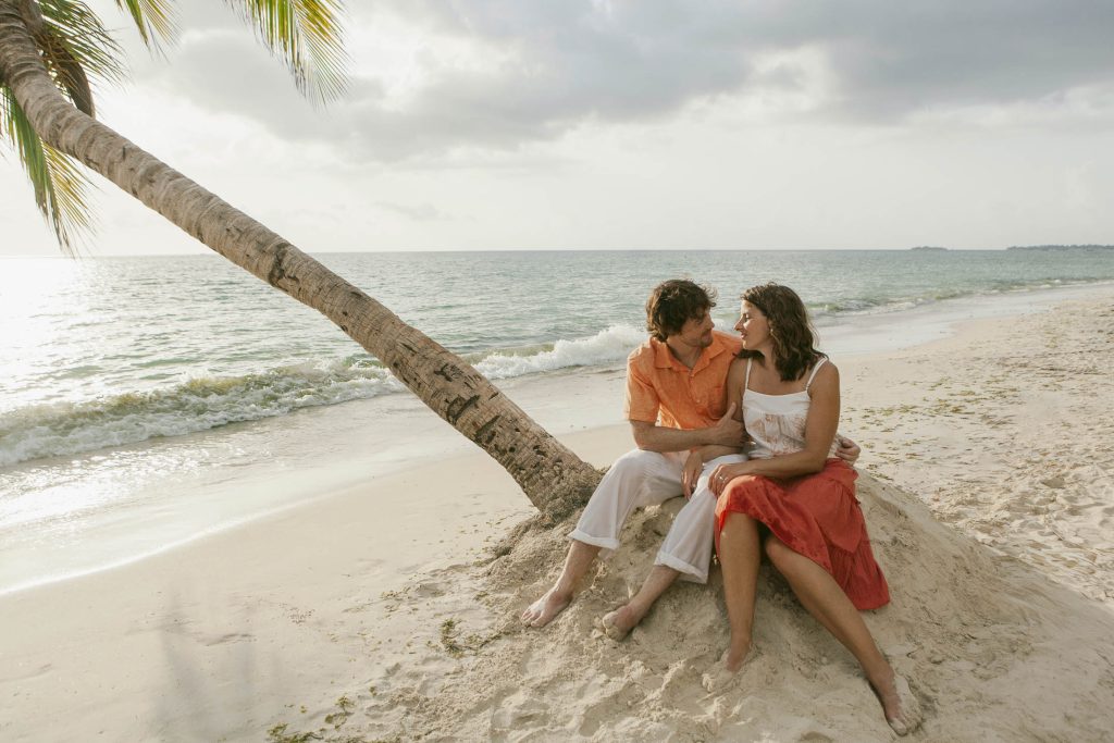 Negril, Jamaica: A couple sits close together on sandy beach near a leaning palm tree, smiling at each other with gentle waves and a cloudy sky in the background.