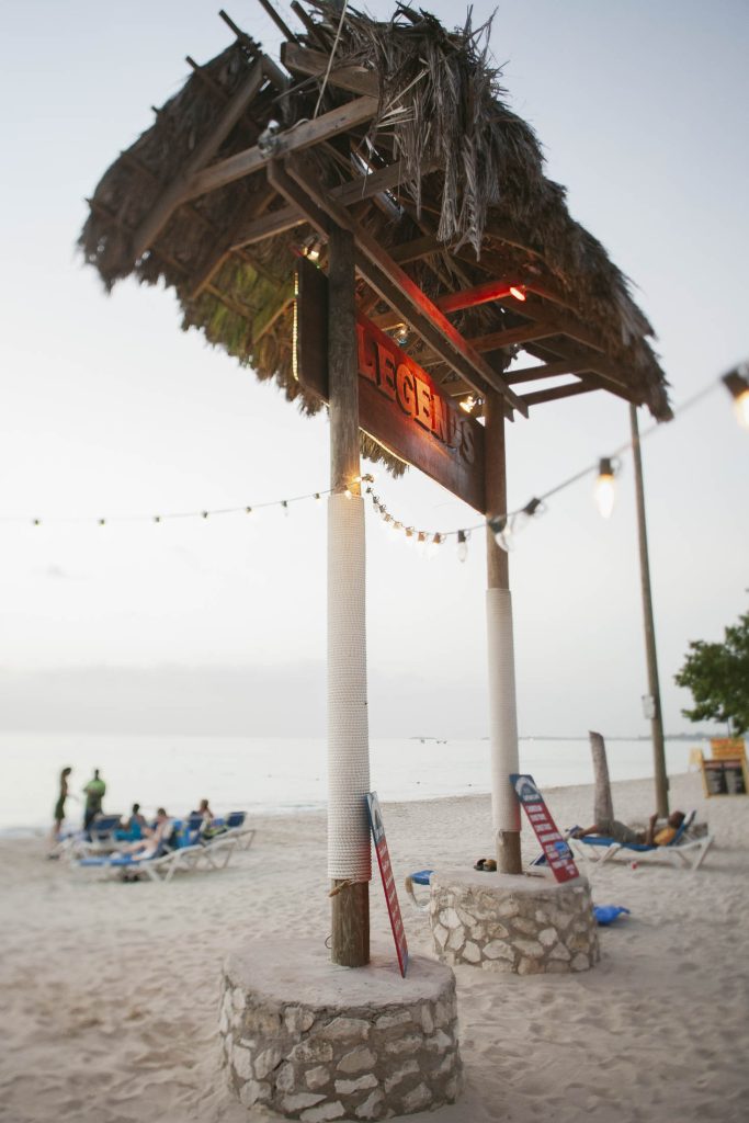Negril, Jamaica: A rustic beachside sign with a thatched roof reads "LEGENDS." String lights hang overhead, and people lounge on beach chairs near the ocean under a cloudy sky.