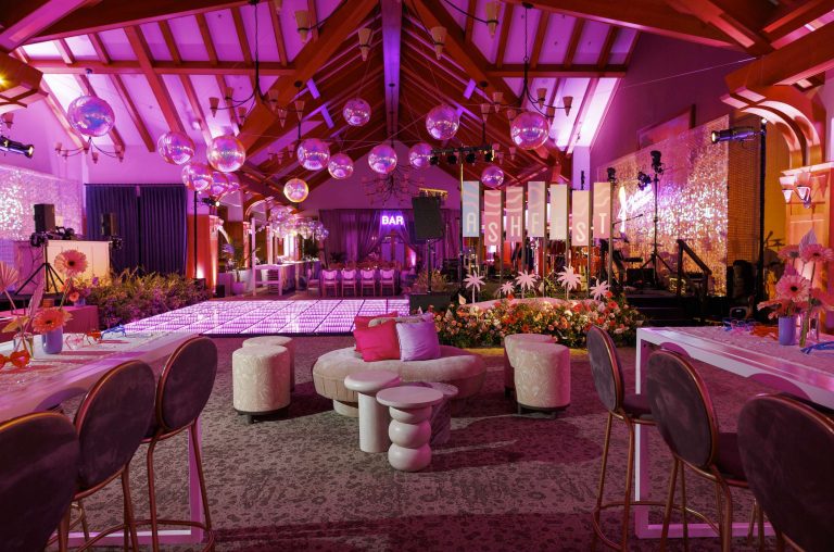 A vibrant event space decorated with pink and purple lighting, disco balls, floral arrangements, elegant seating, and a dance floor. Large letters spell out "SHE IS" near the stage, creating a festive atmosphere.