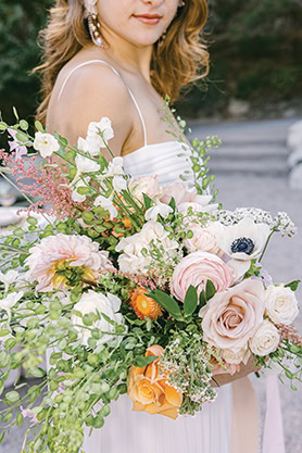 A woman in a white dress holds a large bouquet of assorted flowers, including roses, greenery, and small white blooms. The background is outdoors with greenery. Photographer specializes in Documentary Editorial Wedding Lifestyle